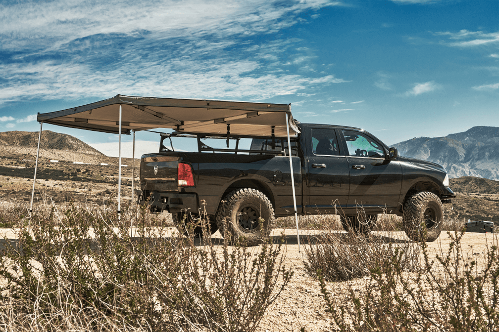 Black pickup truck with a gray 270-degree awning deployed off a bed rack, parked in the desert