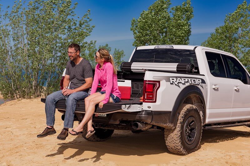 Two people sitting on the dropped tailgate of a white pickup truck parked on sand near the water