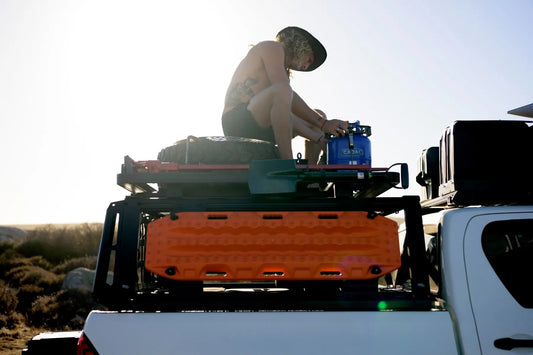 Overlander loading recovery boards and a spare tire onto a black bed rack on a white pickup truck