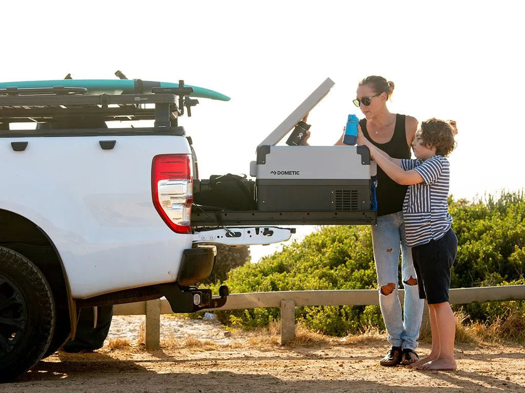 Woman and child opening a portable fridge on a cargo slide pulled out from the tailgate of a white pickup at the beach